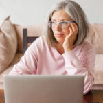 Woman with gray hair and glasses sitting forlorn in front of computer